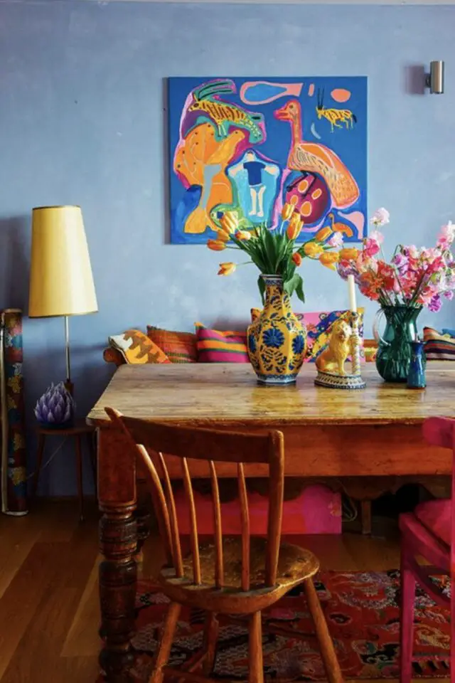intérieur style maximaliste design salle à manger mur bleu table et chaise en bois banquette avec coussin coloré tableau peint vases jaune et vert tapis à fleurs